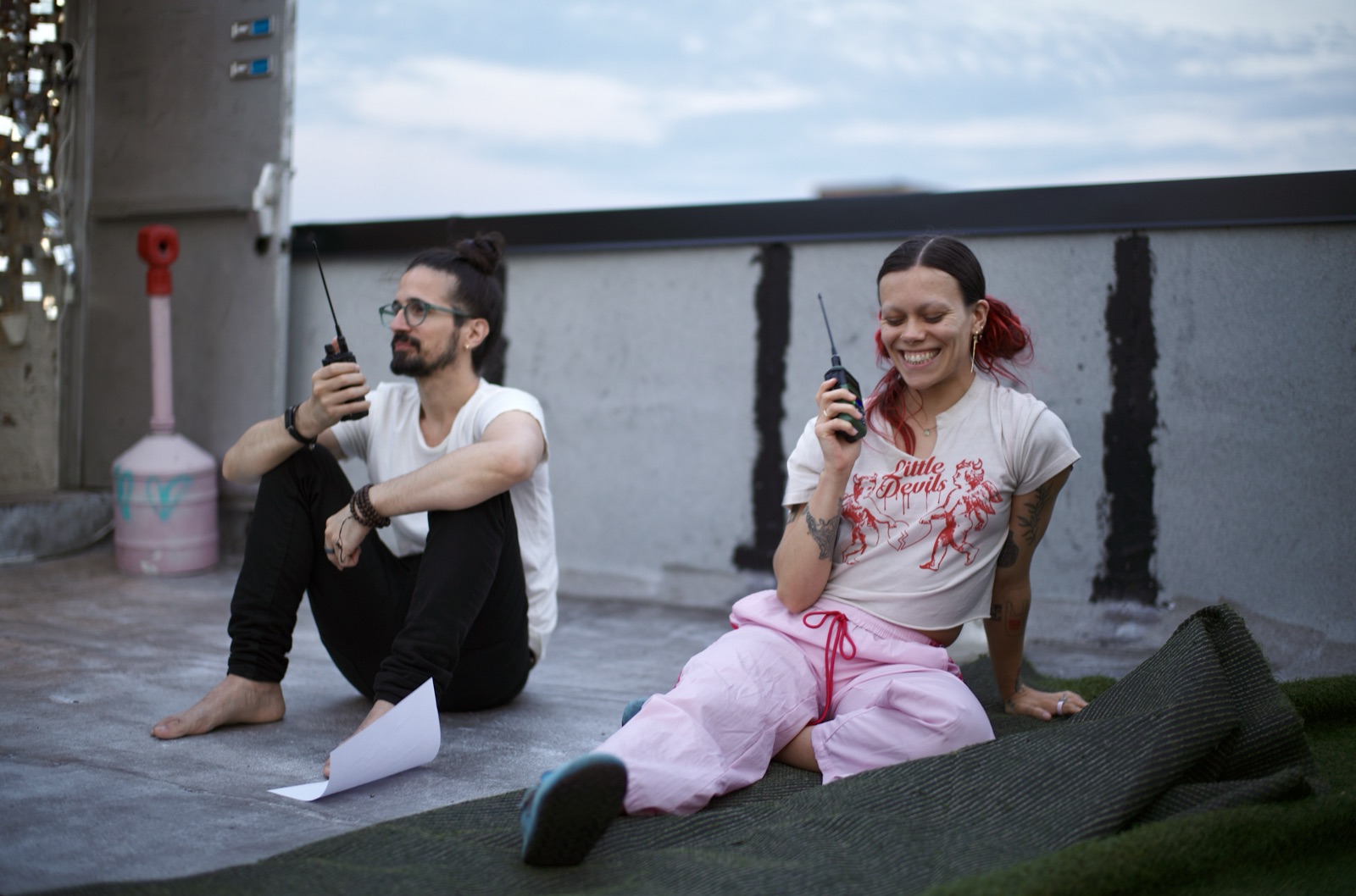 Two participants laugh as they raise their handheld radios on the rooftop turf at dusk.