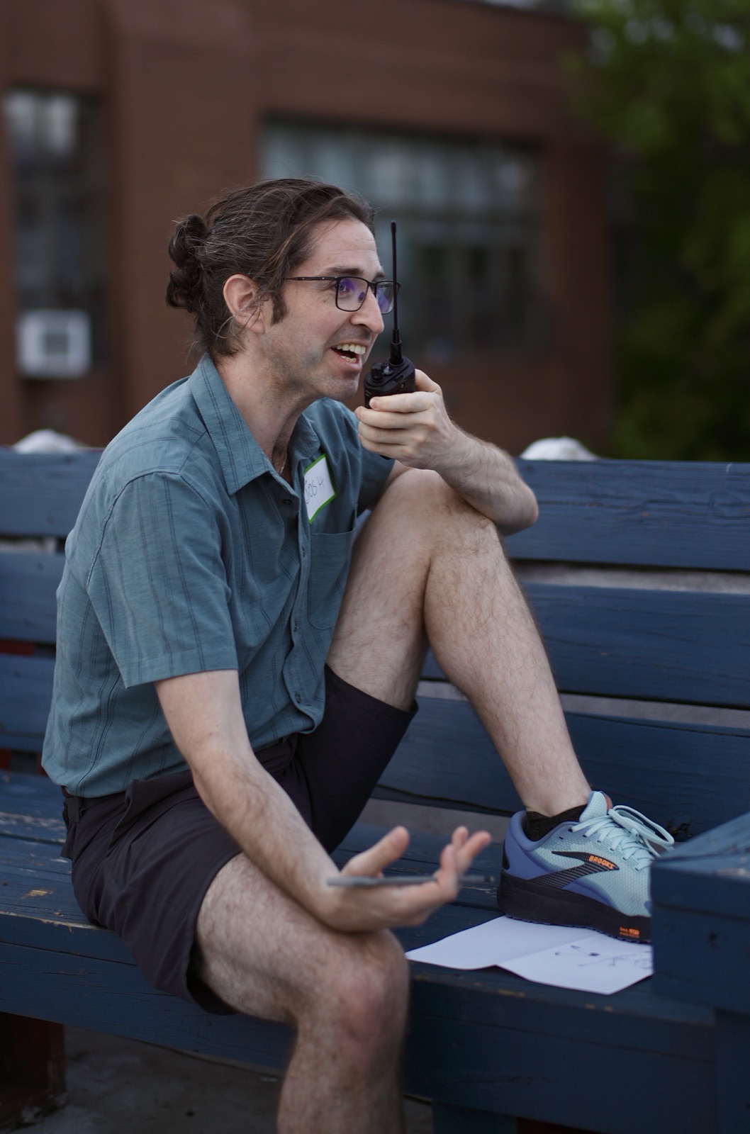 An attendee talks into a handheld radio on a blue rooftop bench, a sheet of paper from the relay game beside him.