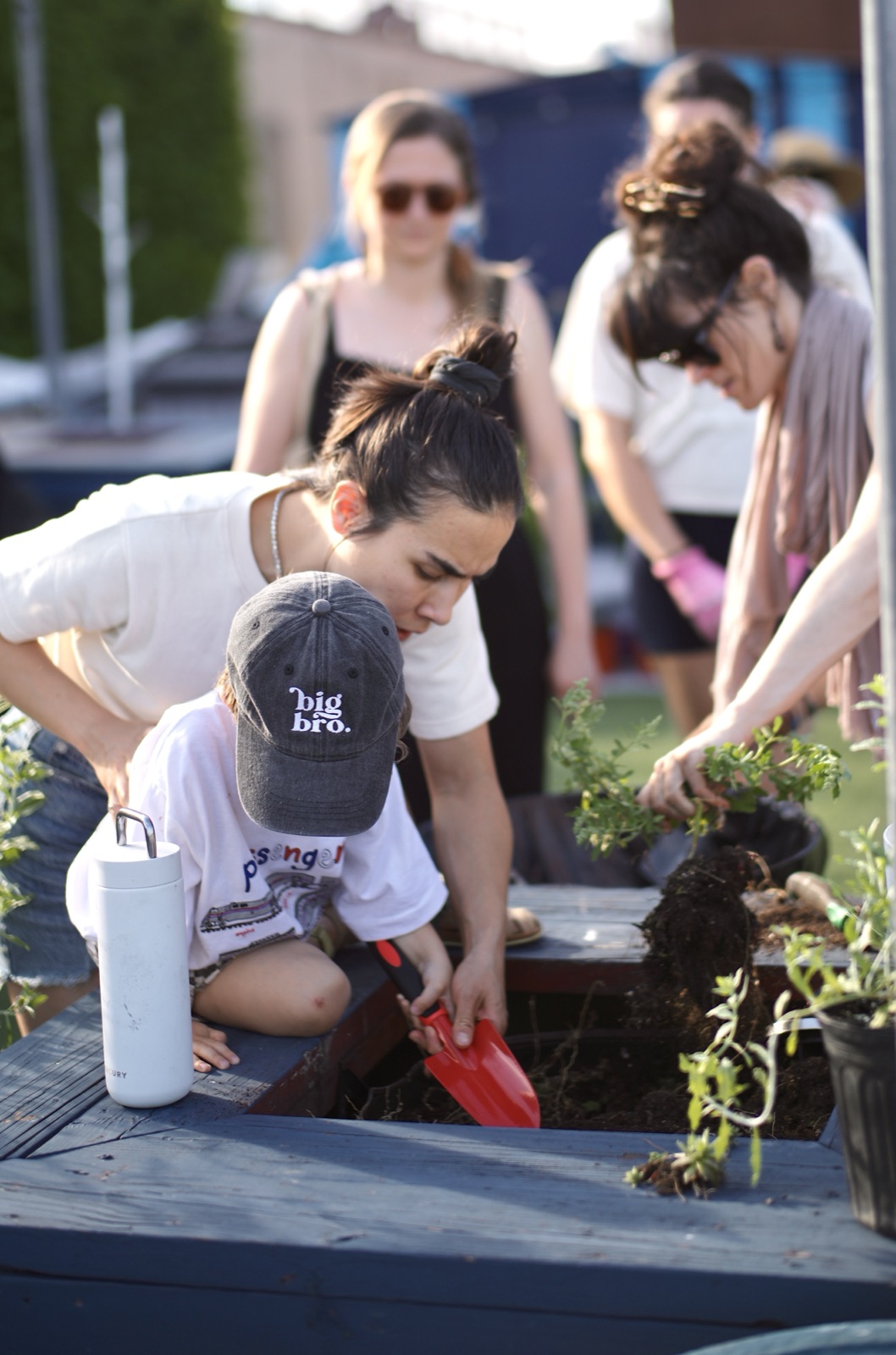 An attendee kneels at a wooden planter, trowel in hand, working soil around a new herb.