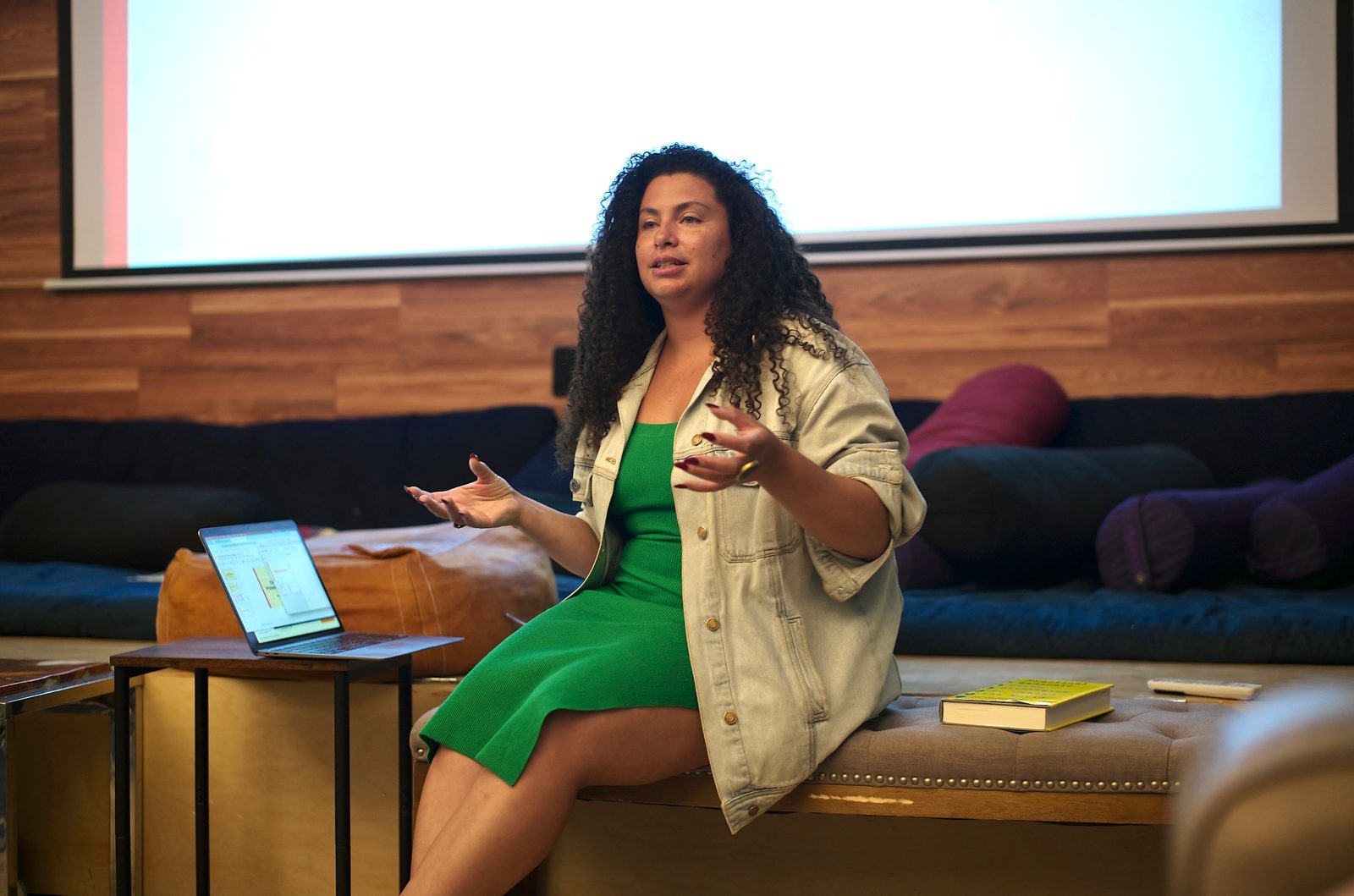 Allison King mid-gesture on the tufted bench, slides on the laptop beside her and a copy of Practical Radicals at hand.