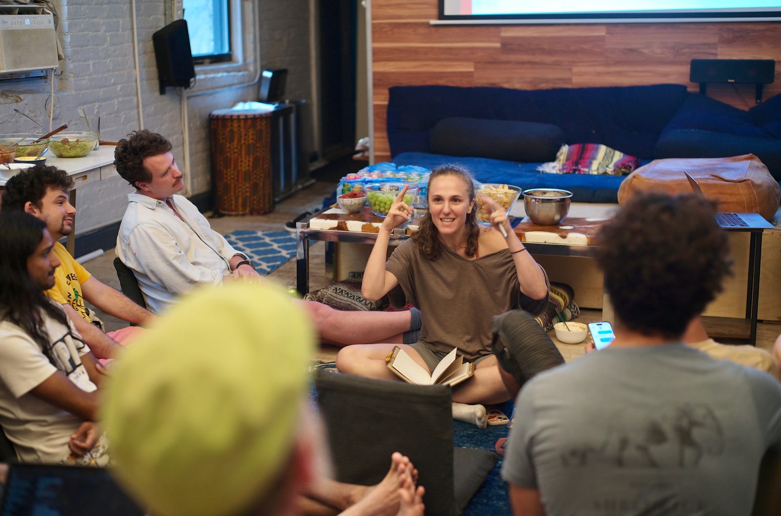 An attendee on the rug, notebook open in her lap, makes a point with both hands raised while the circle turns to listen.