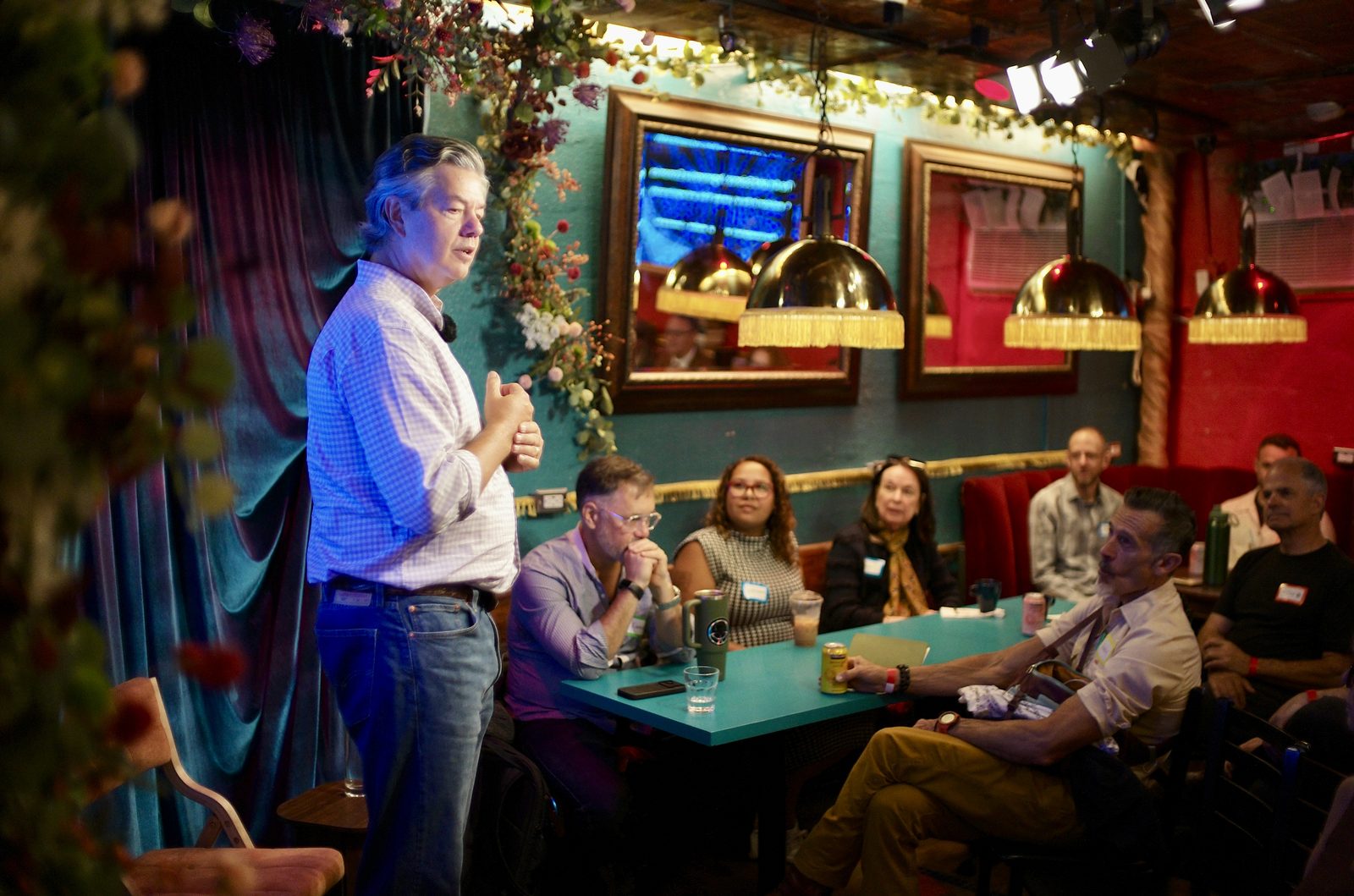 John Fullerton mid-sentence at the front of the back room, hands together, the first row of booths listening under the brass lamps.