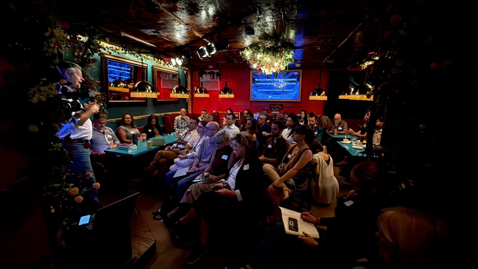 The back room from the side, every booth and chair taken, the title slide lit on the screens at the far end.