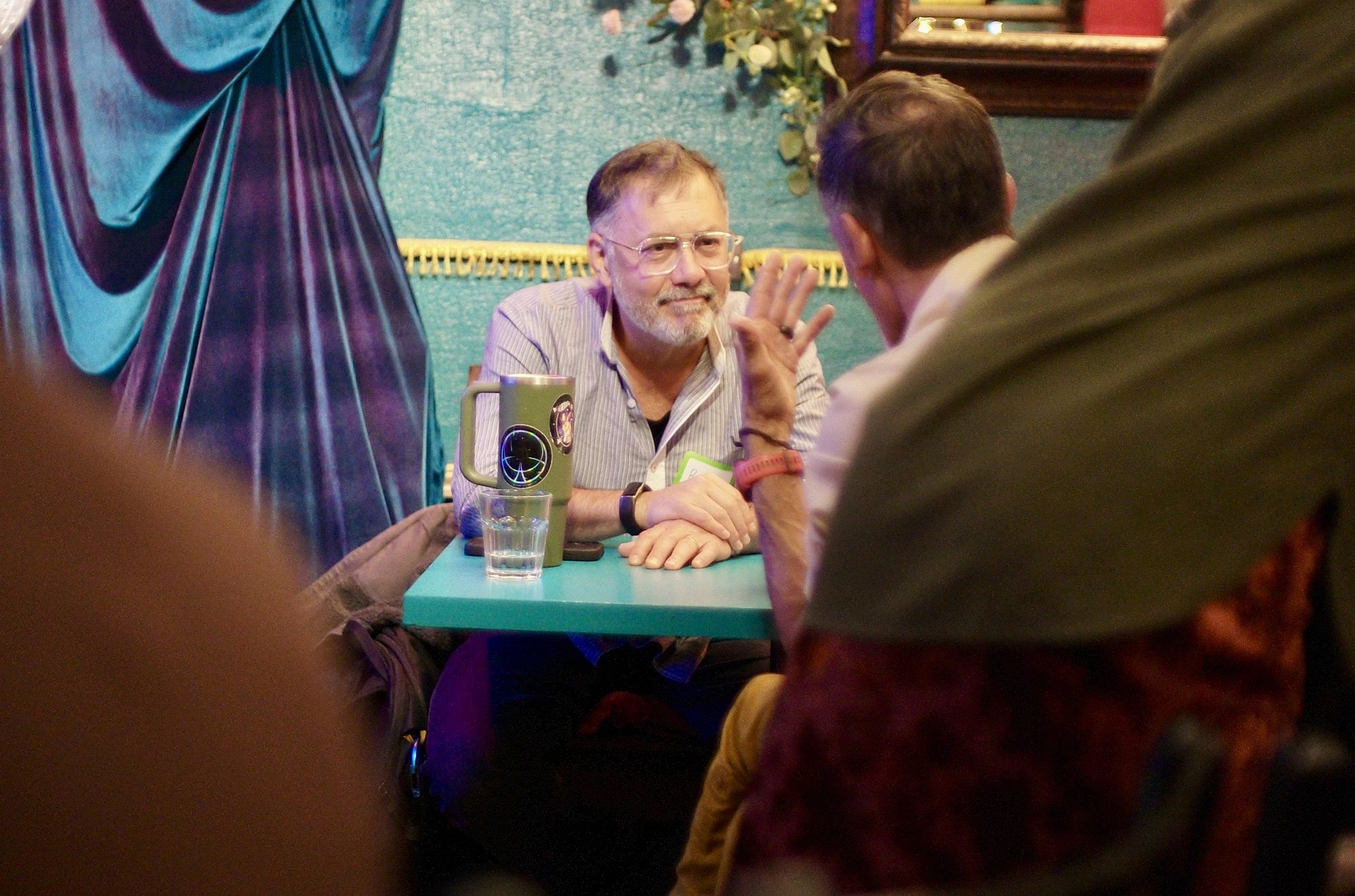 Two people at a booth table during the break, one listening with a half smile, the other mid-answer with a hand raised, working through the one question Fullerton set: how does that story land in your solar plexus?