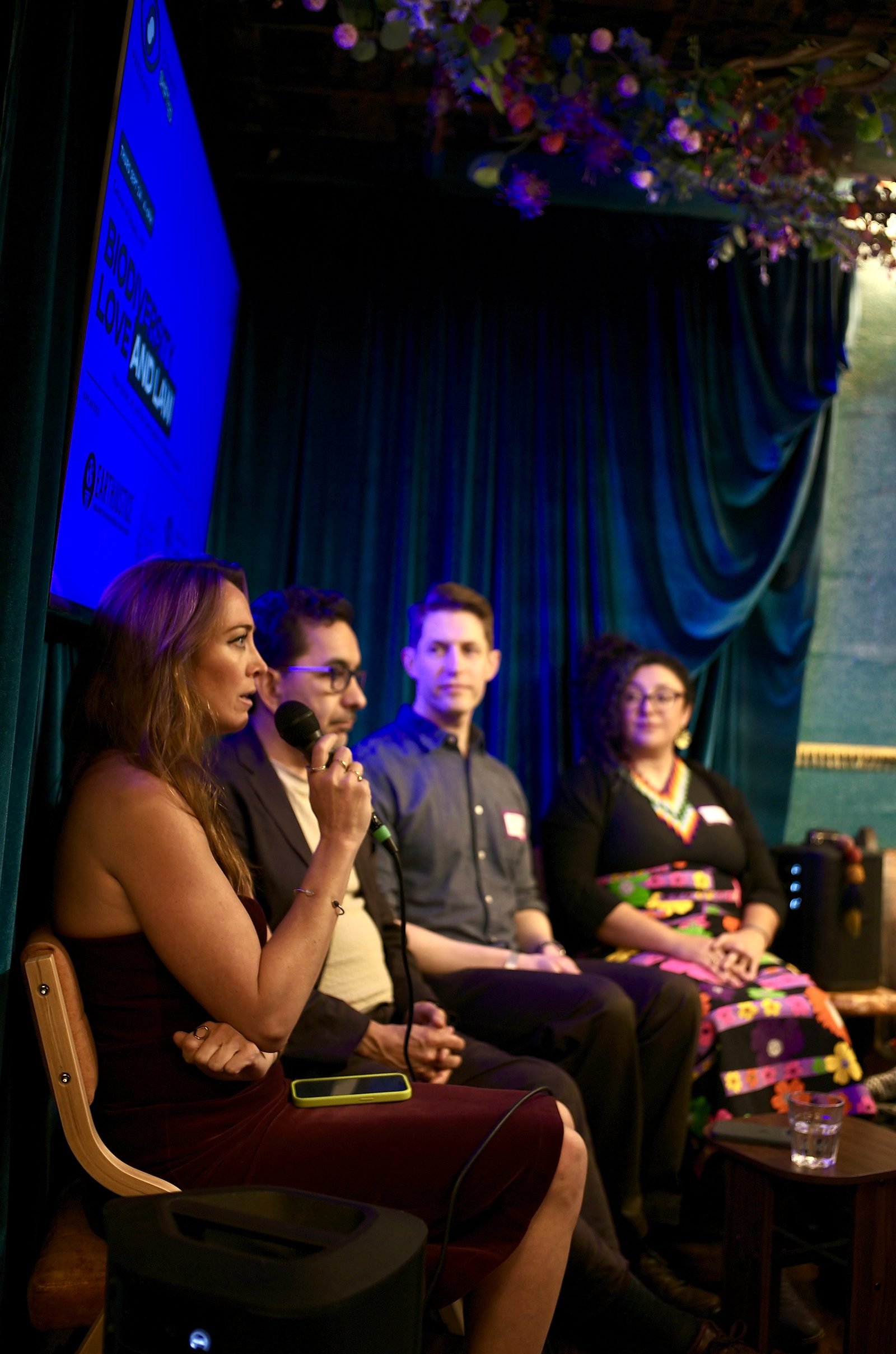 Eva Frye speaking into the microphone at the end of the panel, the title screen glowing behind her.