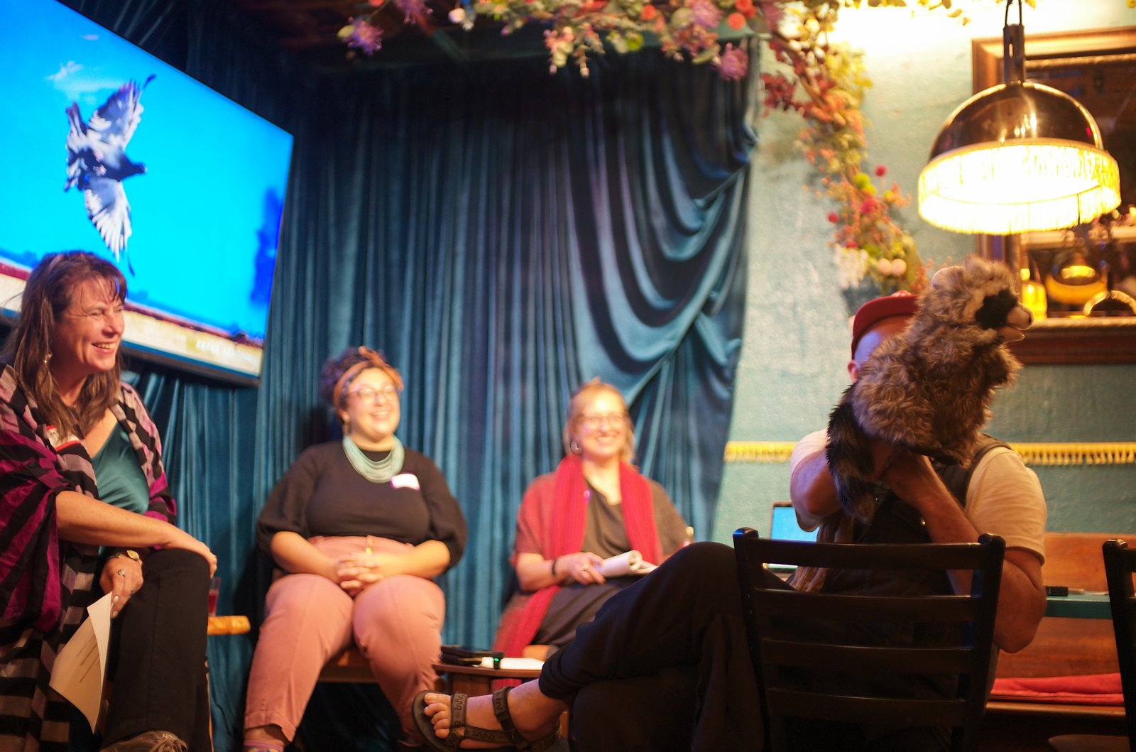 Leah Lamb, Roxi Shohadaee and Mariette Papic laughing on the stage while a man in a red cap holds up a raccoon puppet, a pigeon in flight on the screen behind them.