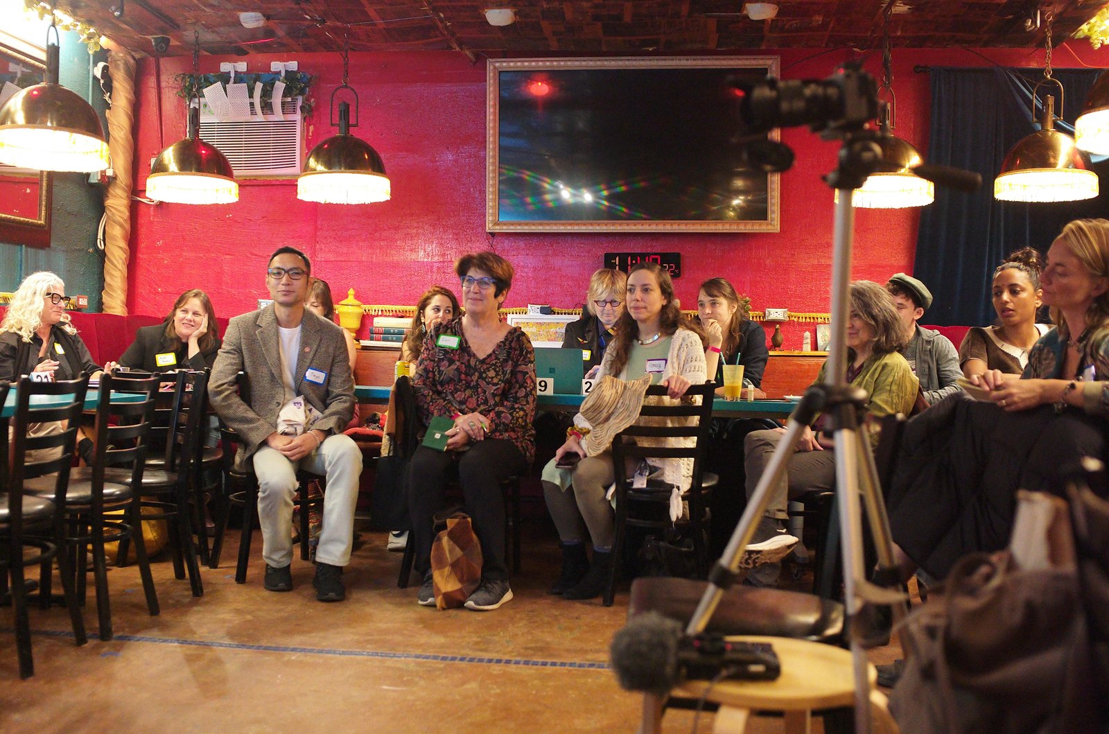 The room listening in a half circle under the brass lamps and the red wall, name tags on, a camera on a tripod in the foreground.