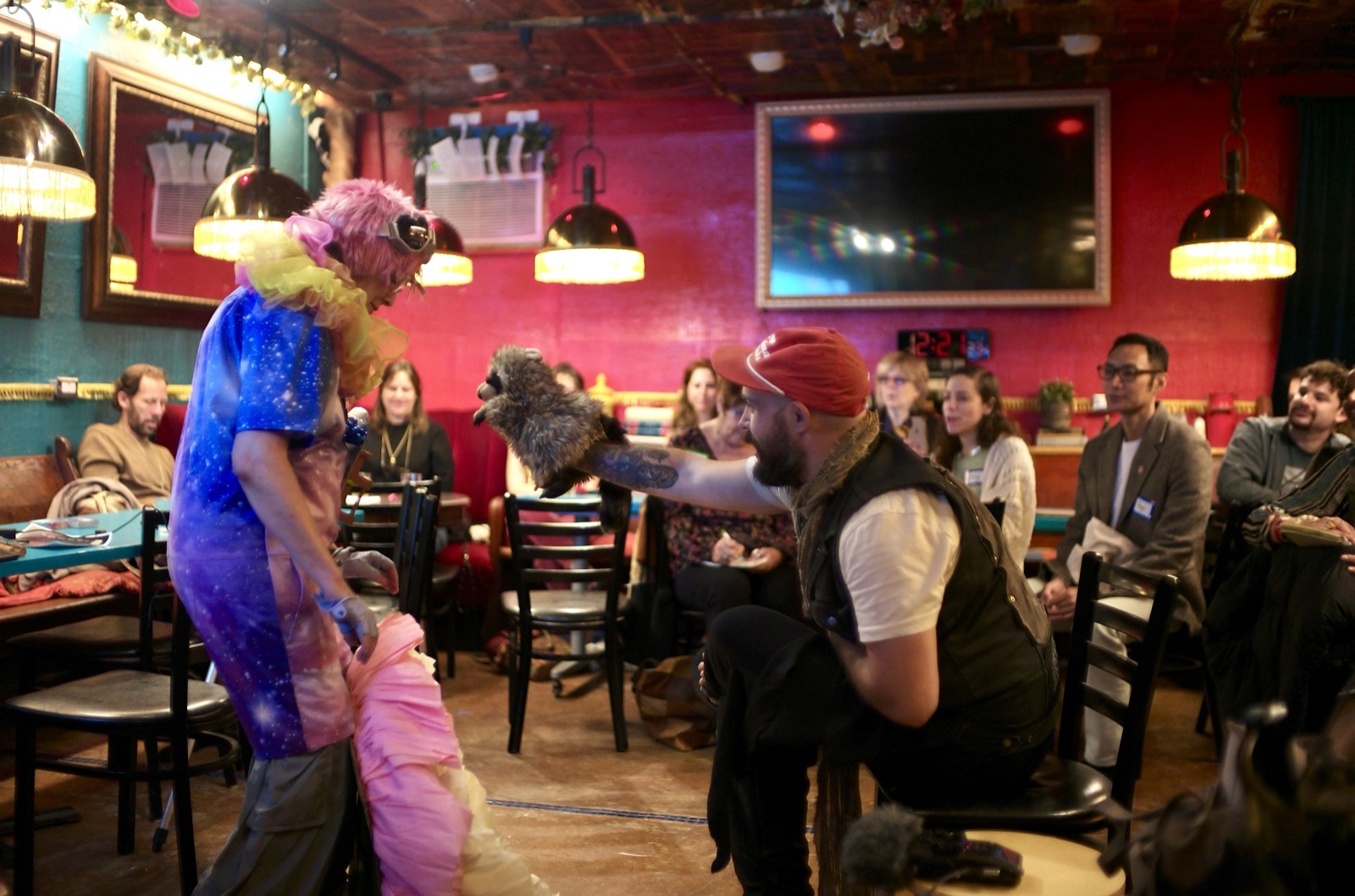 A performer in a galaxy-print costume, a shaggy pink headpiece with goggles and a yellow ruff, turns toward a man in a red cap who leans out from his chair with a raccoon puppet held at arm's length, the room watching from the booths under the brass lamps and the red wall.
