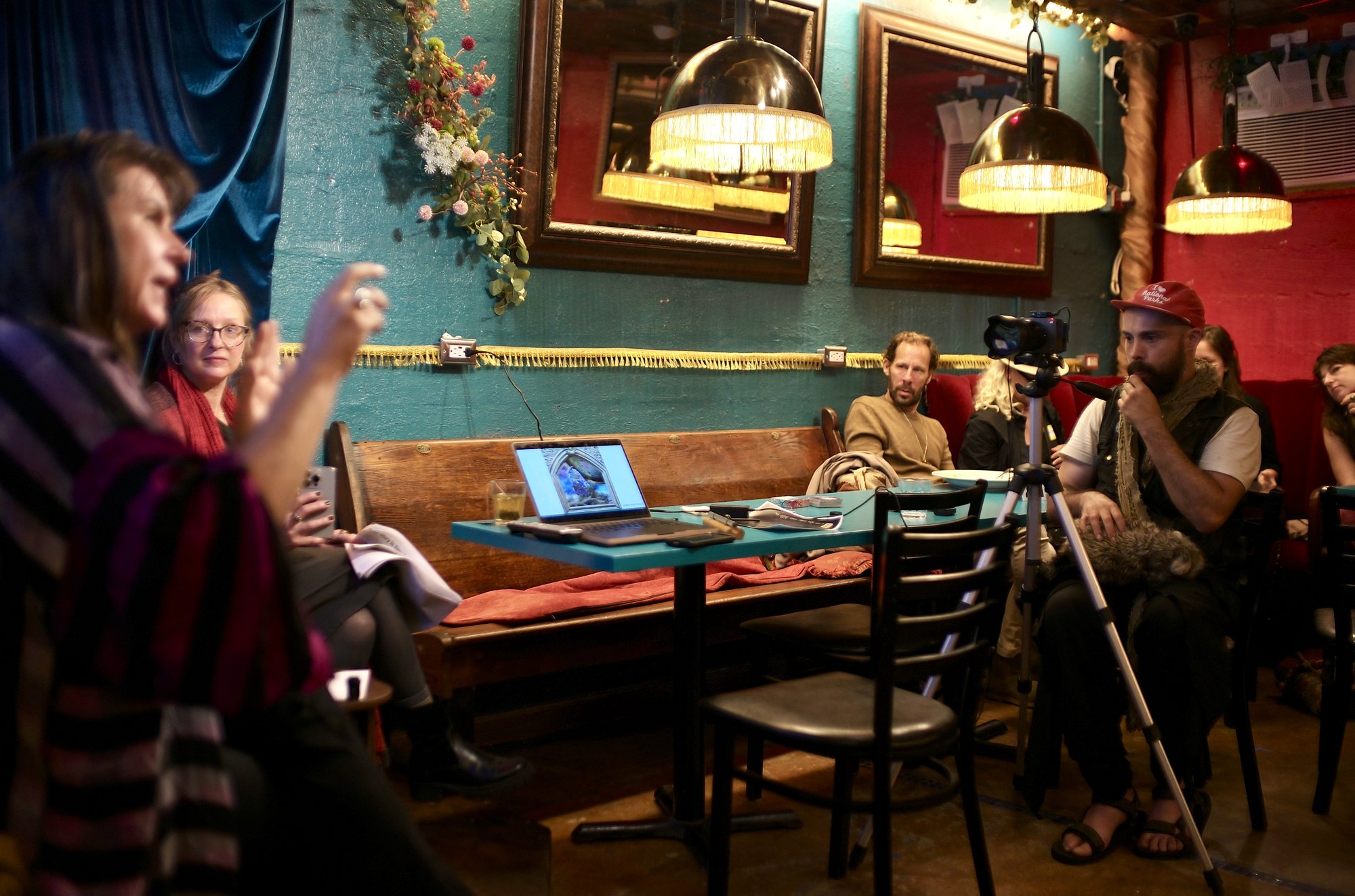 Leah Lamb telling the story of Earth, one hand raised, Mariette Papic beside her and listeners along the teal wall under the brass lamps, a laptop on the table showing an owl in an arched frame.