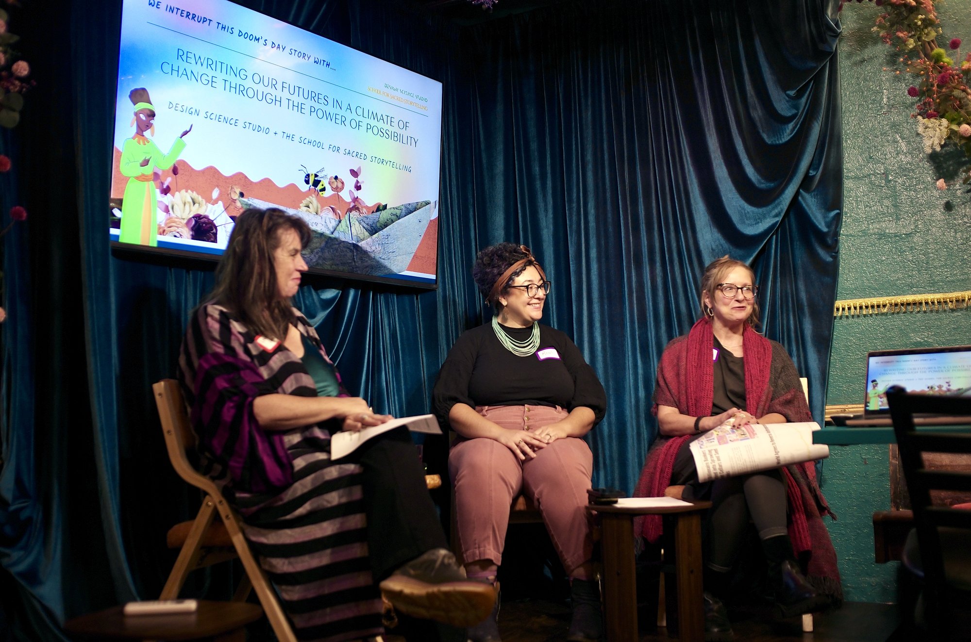Leah Lamb, Roxi Shohadaee and Mariette Papic, left to right, seated before the blue velvet curtain, Papic holding a rolled newspaper. The title slide on the screen above them reads: We interrupt this doom's day story with... Rewriting our futures in a climate of change through the power of possibility. Design Science Studio + the School for Sacred Storytelling.