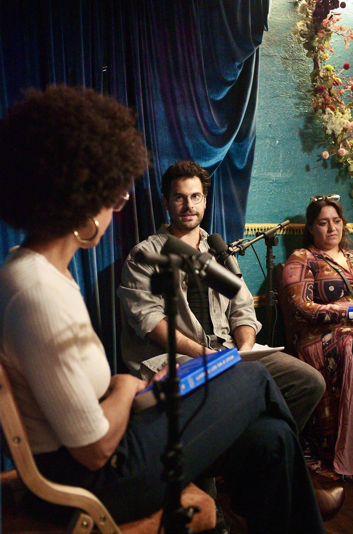 Michael Mezzatesta listening across the microphones, Ayana Elizabeth Johnson's back to the camera with a blue copy of her book on her knee, a producer watching from the side.