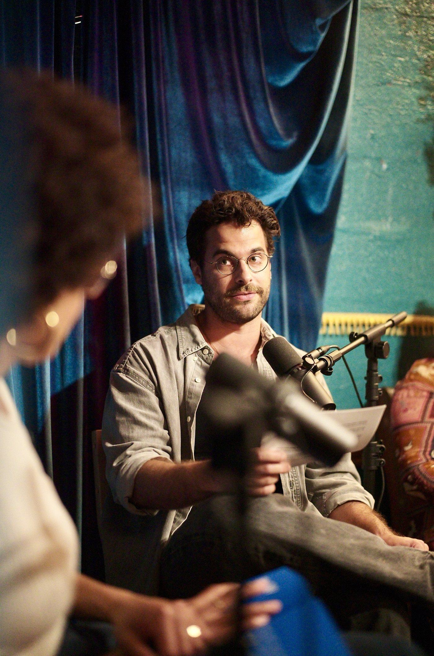 Mezzatesta at the microphone in front of the blue velvet curtain, notes in hand, mid-question.