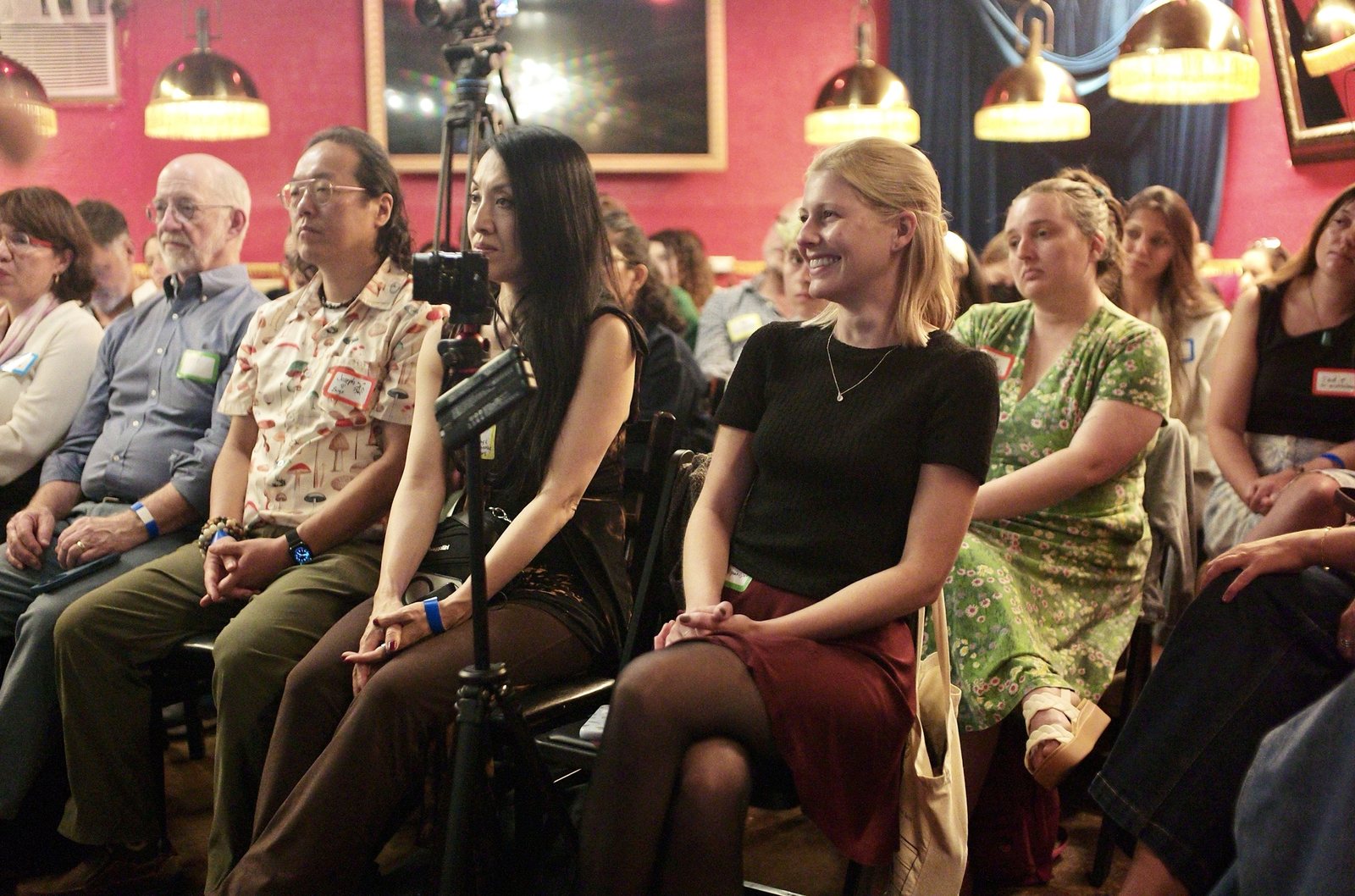 The front row under the brass lamps, a camera on a tripod between the chairs, one listener smiling toward the stage.