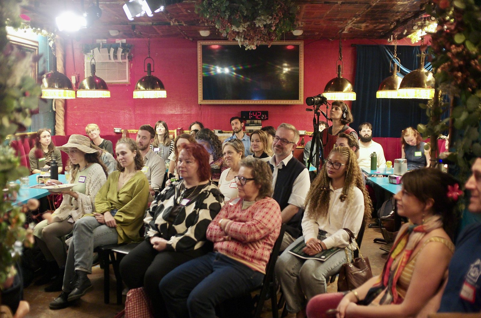 The room before the panel: the banquettes and chairs full under the fringed lamps, most faces turned to the stage.