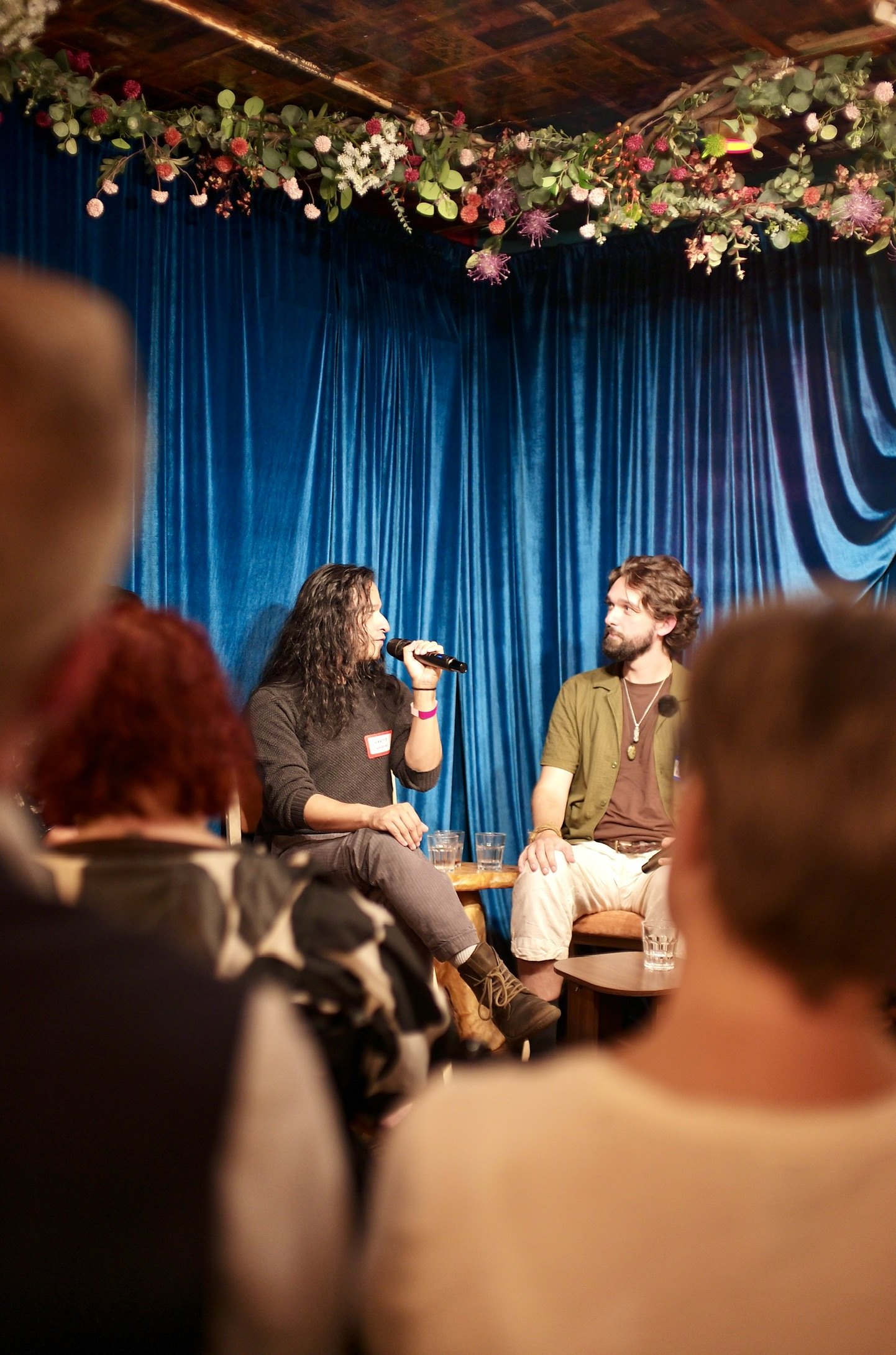 Jeremy D Johnson speaking, Henry Kramer beside him, under the flowers over the curtain.