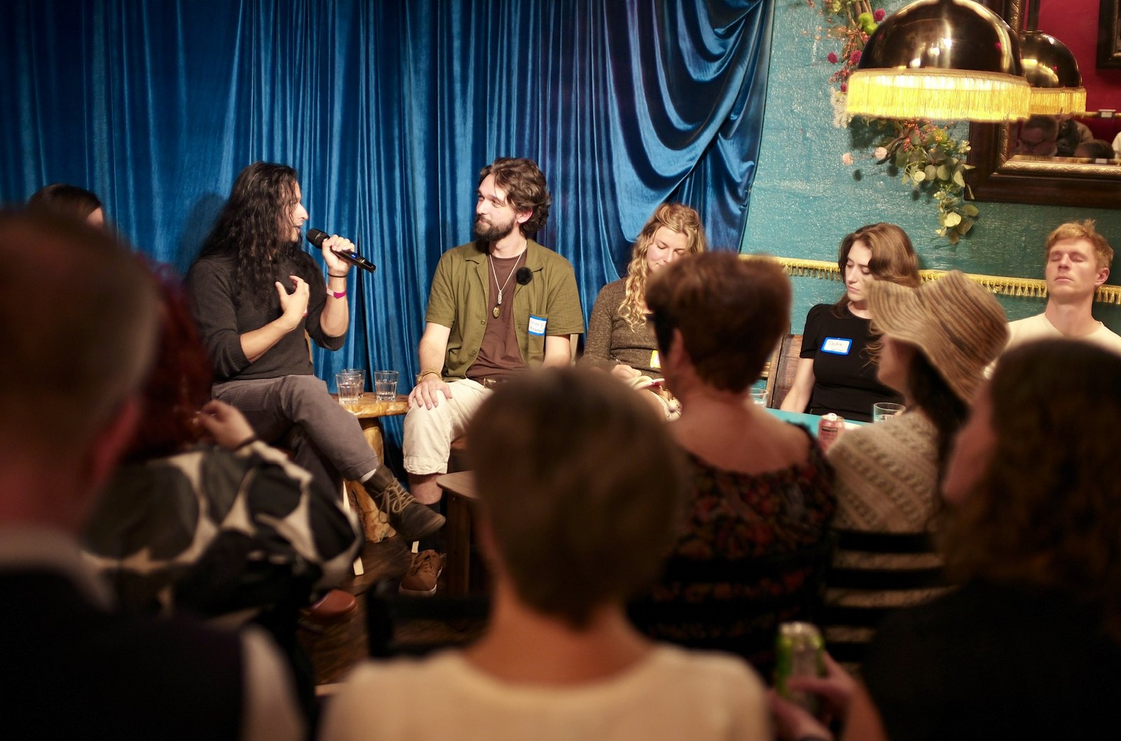 The panel from the back row: five heads in the foreground, the stage lit under the brass lamp.