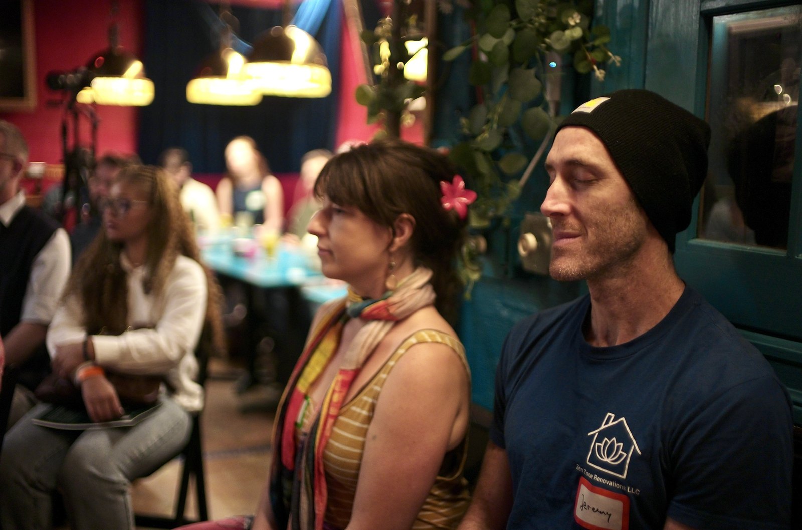 Two listeners at the side of the room, one in a knit cap, both looking toward the stage.