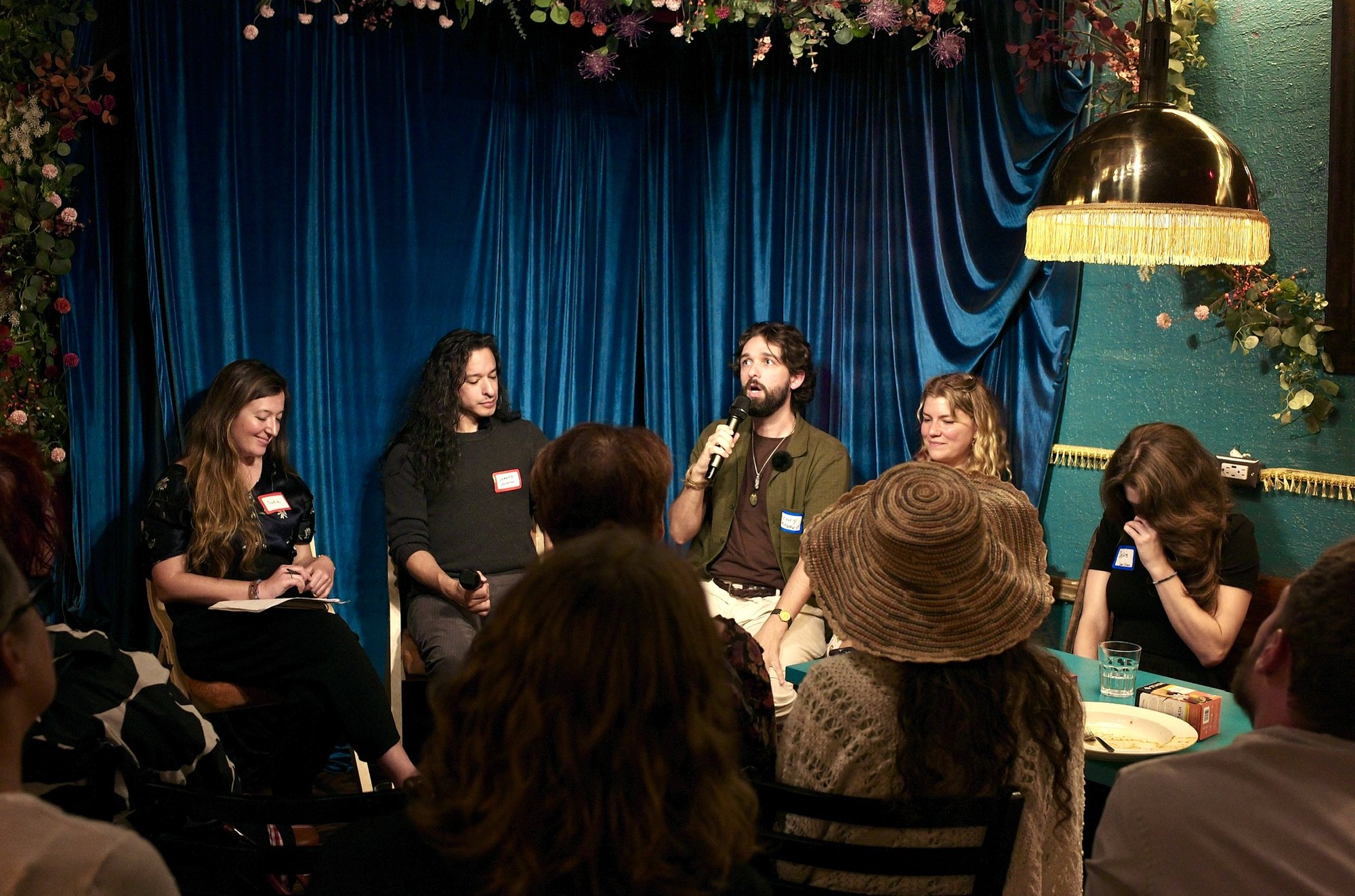Henry Kramer speaking under the brass lamp, the other three panelists listening along the curtain.