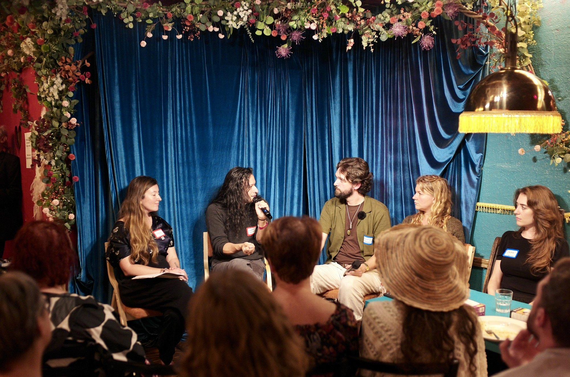 The panel under the flowers and the blue curtain: Jeremy D Johnson speaking into the microphone, Henry Kramer turned toward him, Sadie Rittman with her notes at left, the brass lamp overhead.