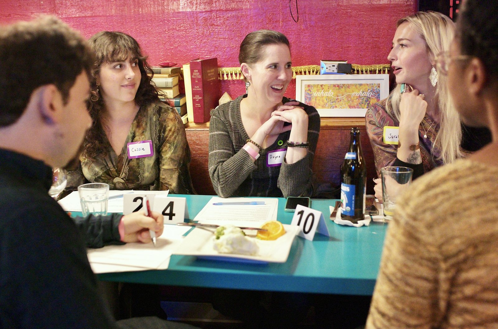 A table under the red wall laughing mid-conversation, name tags on, worksheets and table numbers in front of them.