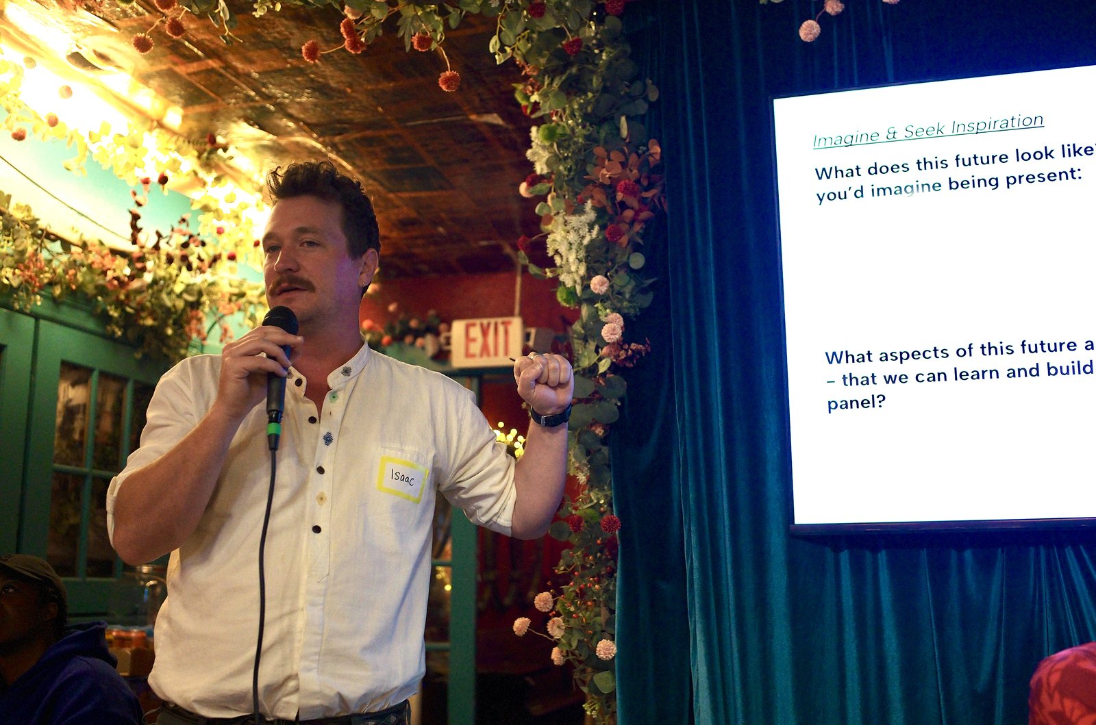 A participant at the microphone reporting back, one fist raised, beside the slide asking what this future looks like.