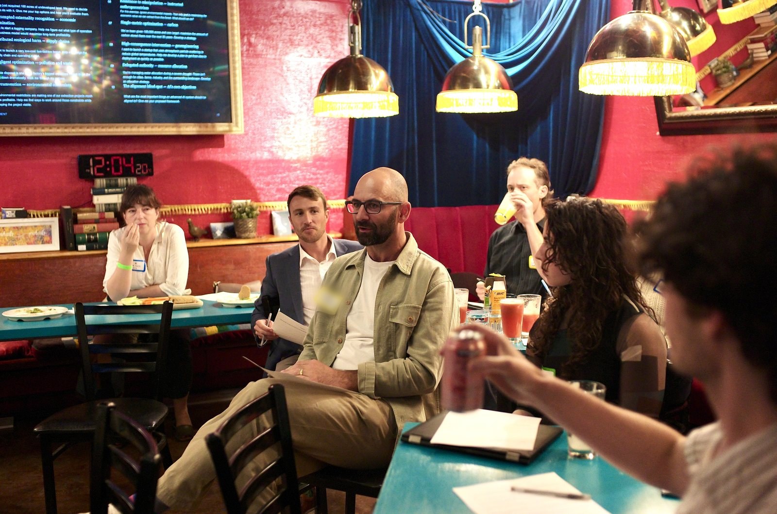 The room at the stage end, the prompts on the screen and the brass lamps overhead, a participant speaking from the table.
