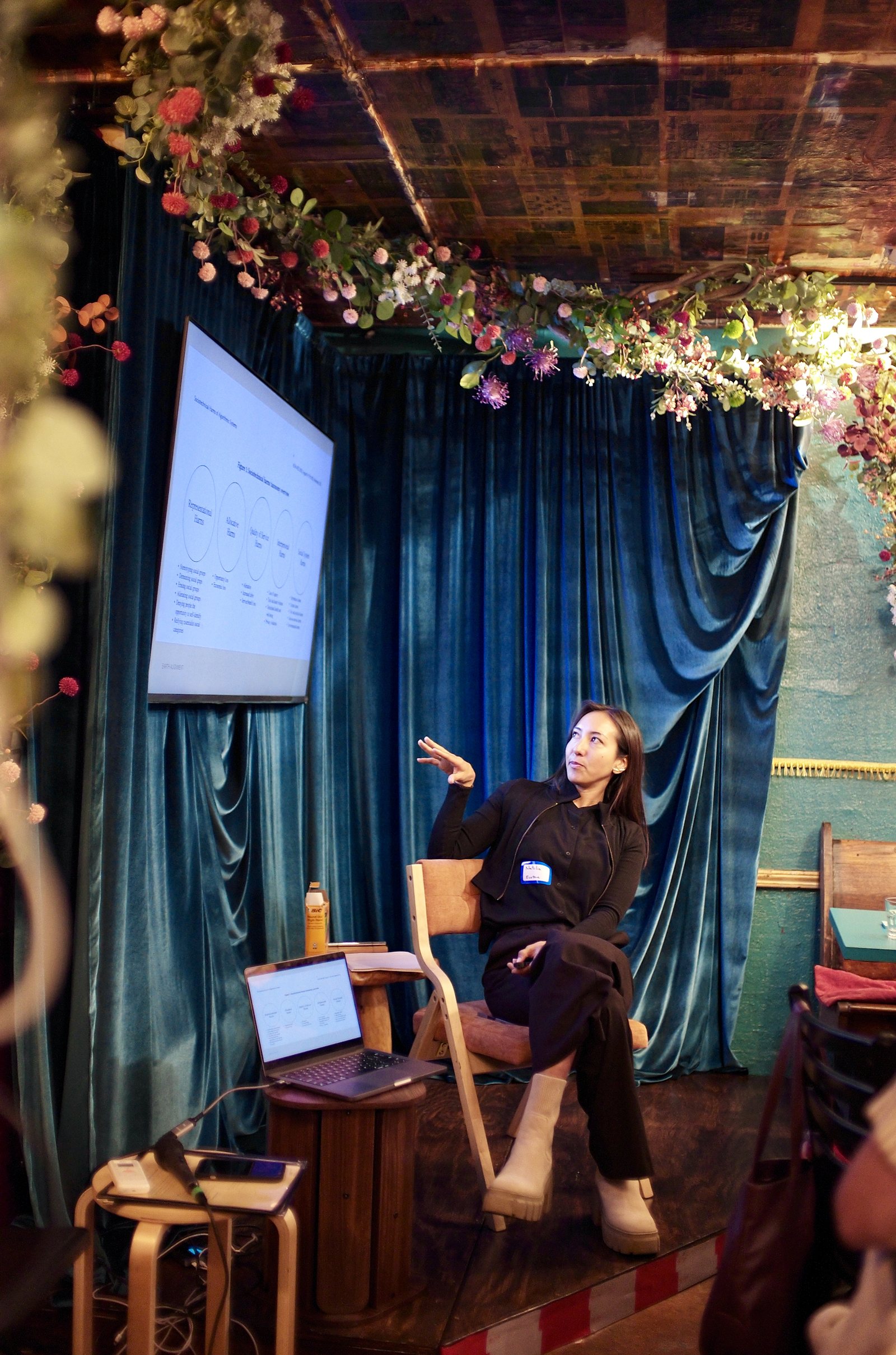 Natalia Vasquez on the stage chair under the flowers, one hand raised, the slide on the screen beside her.