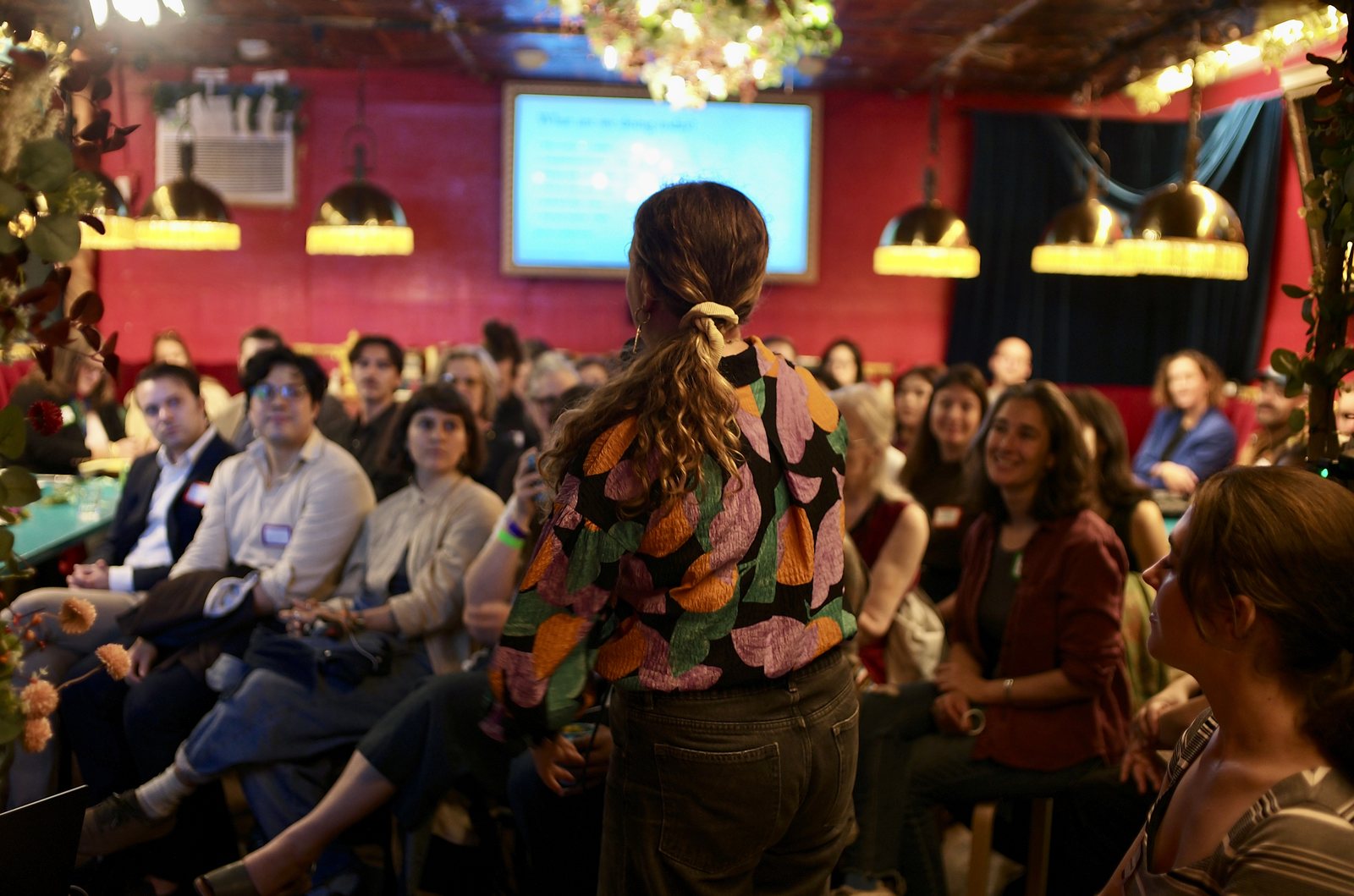 The room from behind a host standing at the front, every seat taken, the agenda on the screen at the far end.