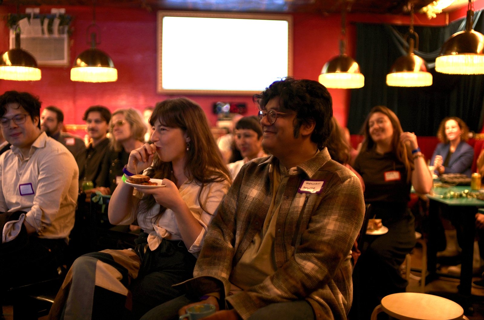 Listeners in the middle rows laughing, one with a plate of food, name tags on.