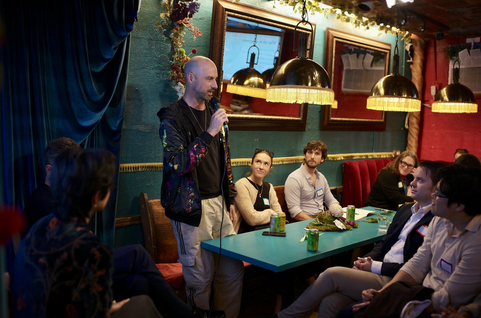 A participant standing with the microphone by the blue curtain, the front table turned to listen.