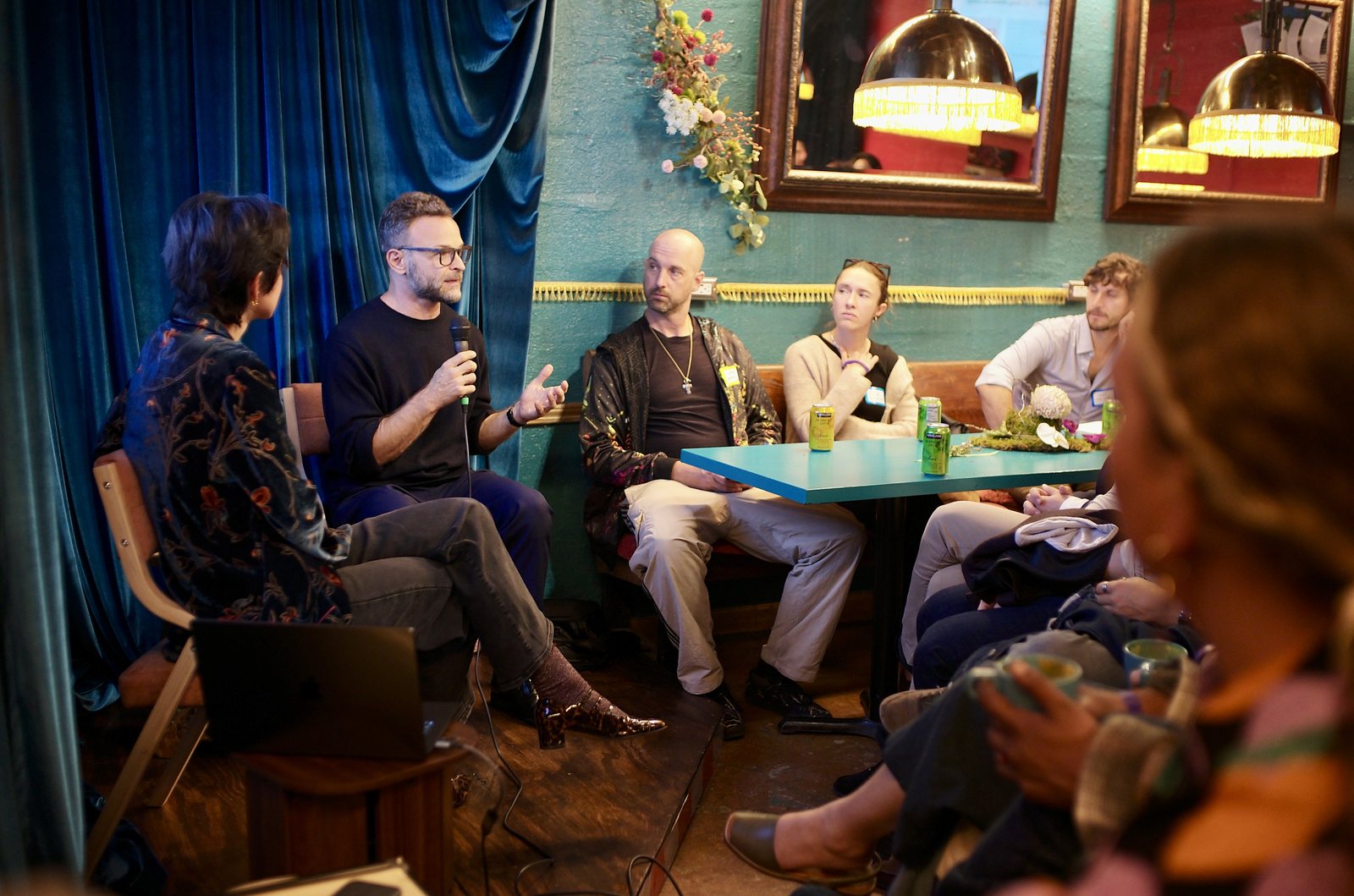 The front table from the audience, a speaker mid-answer with the microphone, seltzer cans on the blue table top.