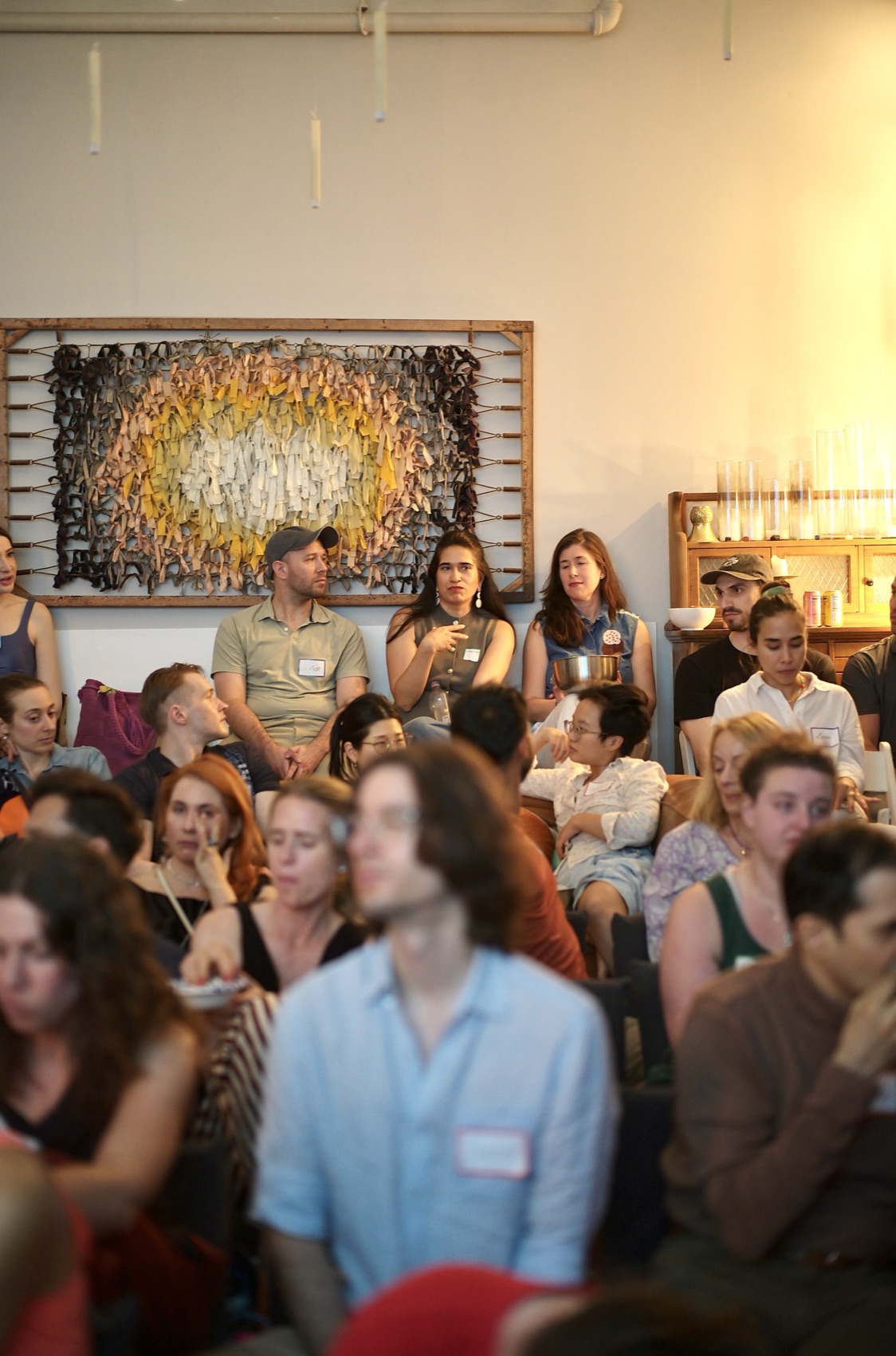 The room at capacity — attendees on chairs and the floor, listening beneath a woven fiber artwork.