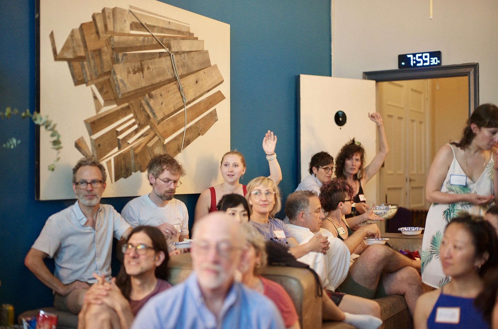 Hands go up across a packed living room as Lent asks who feels collapse is likely — couches, floor cushions, and a wood-assemblage artwork on the wall.