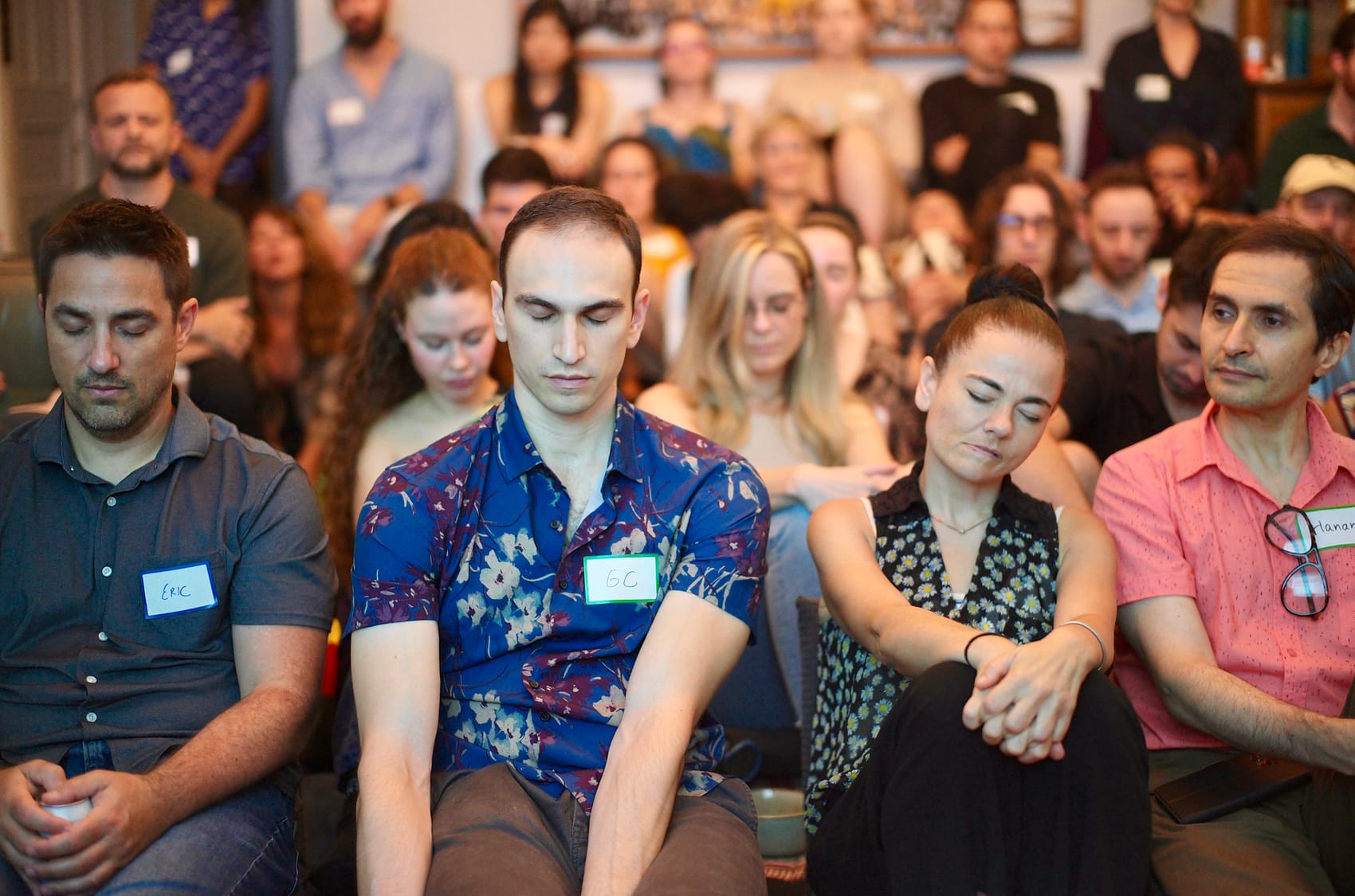 The front rows sit with eyes closed during Hope's opening grounding, nametags on, shoulder to shoulder.