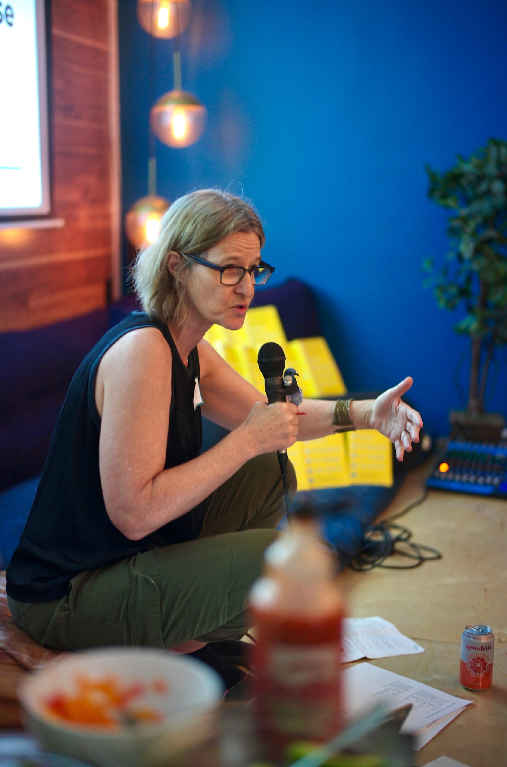 Stephanie Luce speaks from the low stage, mic in one hand and the other mid-gesture, against Lightning Society's blue wall.