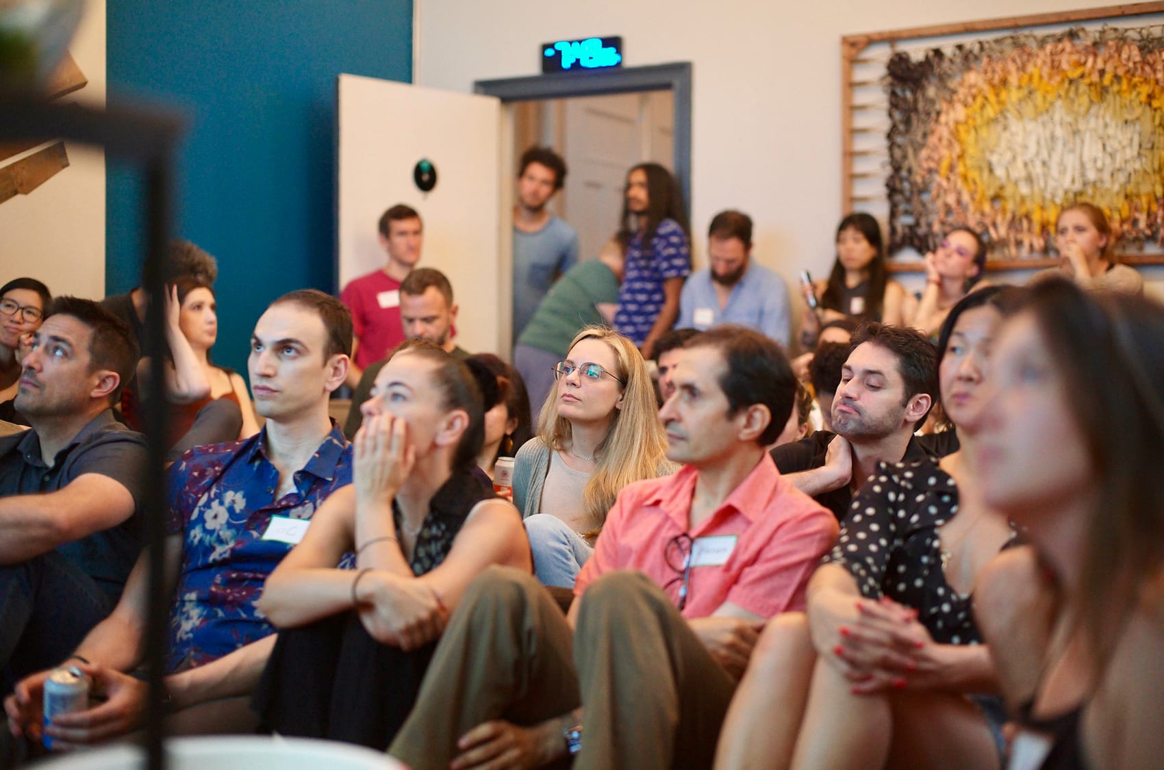 Attendees fill the couches and floor, listening under the woven wall artwork, the clock reading 7:36.
