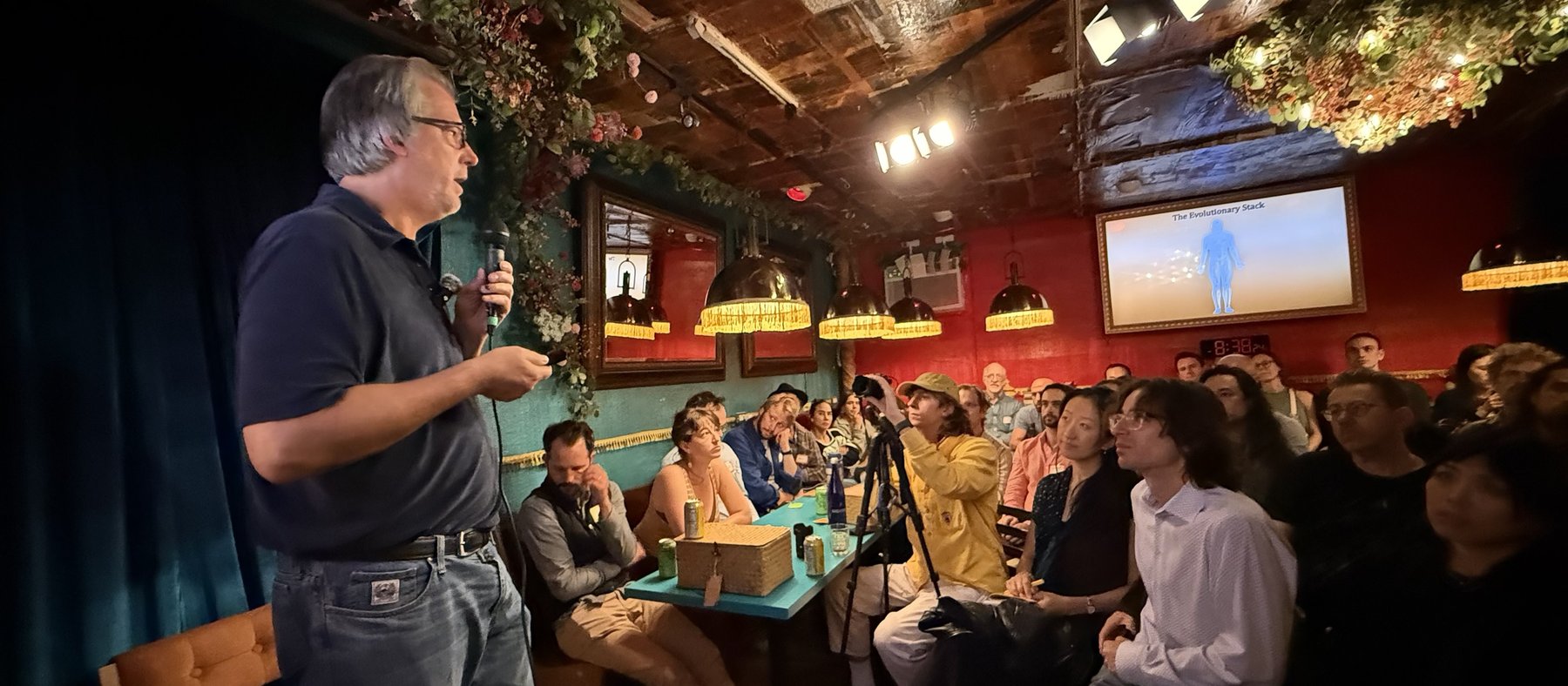 Nate Hagens on stage with the microphone, the room turned toward him and the Evolutionary Stack on the screen behind them.