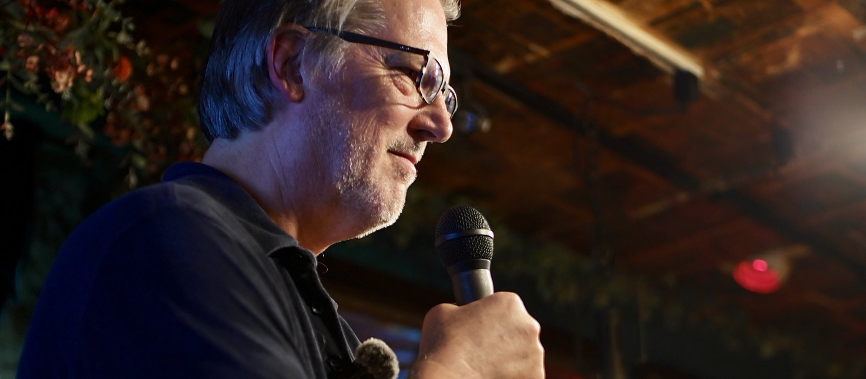 Nate Hagens in profile with the microphone, arms folded, under the pressed-tin ceiling and the flowers of the Dream Room.